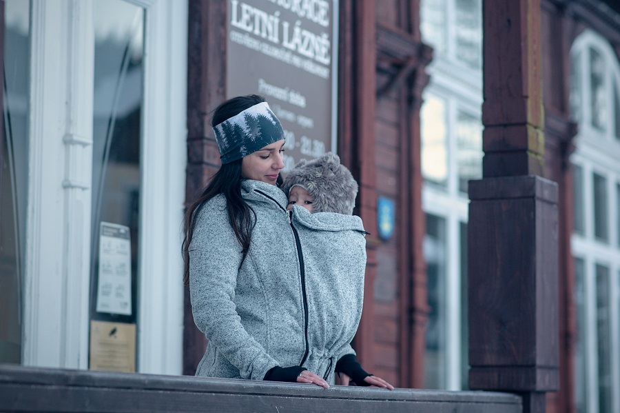 Mladá žena má pod teplou šedou nosící mikinou miminko s beranicí na hlavě
